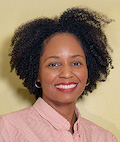 A Black woman with natural hair smiles widely. She wears red lipstick, silver hoop earrings, and a pink striped shirt and stands against a chartreuse wall.