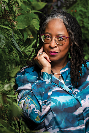 A waist-up photo portrait of Camille Dungy — she wears a patterned blue-and-white shirt and poses thoughtfully against a background of leaves.