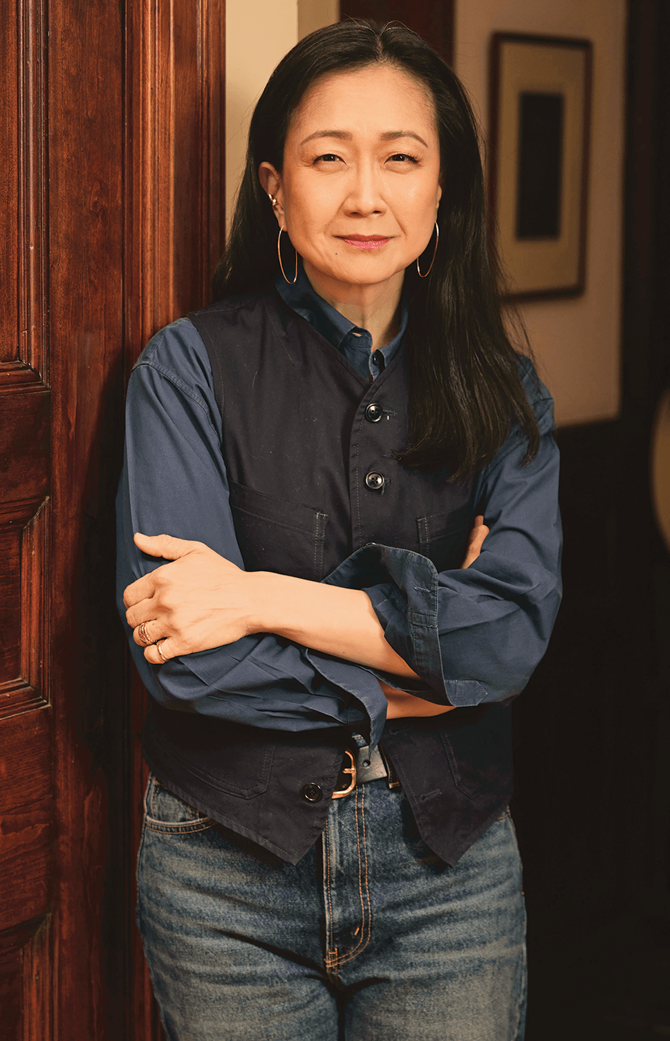 Indoor photo portrait of a woman with long dark hair, hoop earring, and a dark vest over a blue shirt and jeans leans agains a wooden doorframe in a hallway 