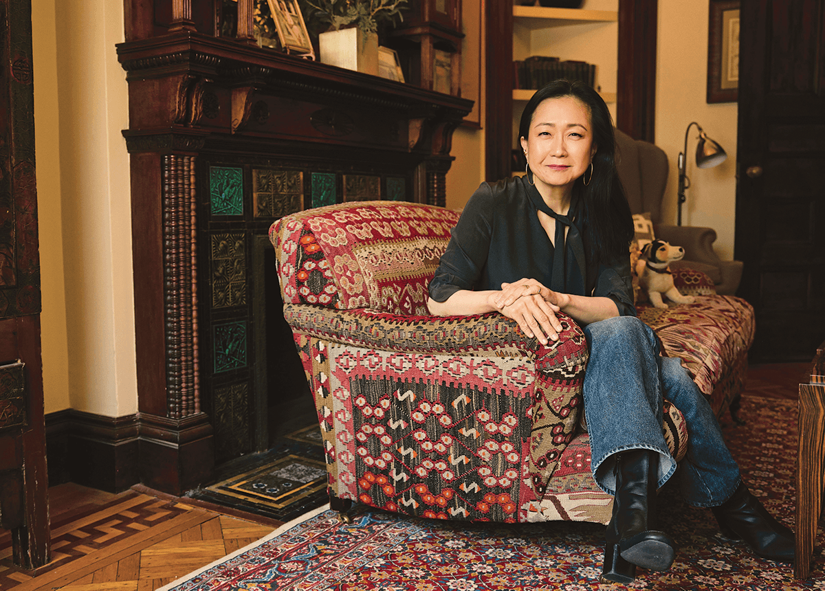 Indoor portrait of a woman with long dark hair, dark blouse, and jeans sits on a printed loveseat in front of a fireplace
