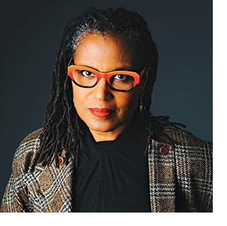 A photo portrait of Anita Gail-Jones, a Black woman with orange-rimmed glasses and locs. She wears a brown plaid blazer and a black shirt.