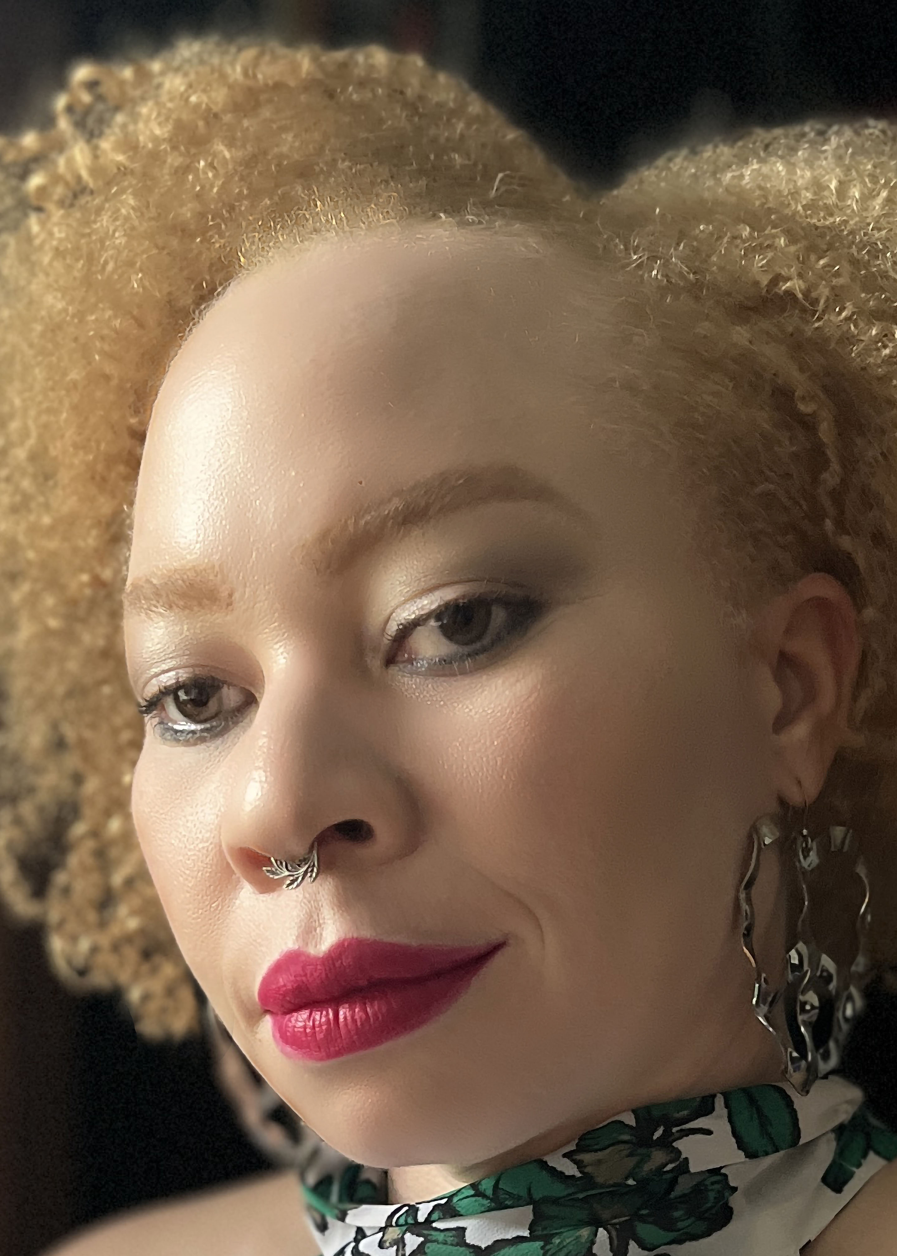 Close-up photo portrait of a woman with parted curly hair, large earrings, a small nose ring, and pink lipstick