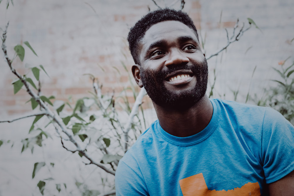 Nigerian American poet Ayokunle Falomo in a light blue t-shirt with short hair and a beard smiles and looks up.