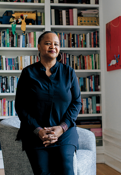 Indoor photo portrait of a woman with her hair pulled back and large hoop earrings leaning on a sofa in a room with many books on shelves
