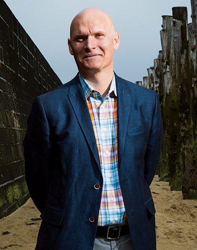 Outdoor photo portrait of a bald man wearing a navy blazer and plaid shirt standing between a stone wall and wooden pylons 