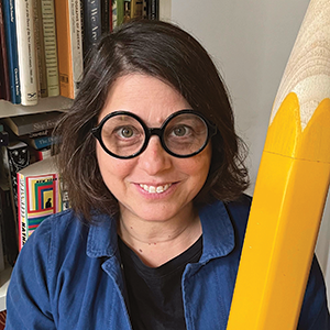 Indoor photo portrait of a woman with round glasses holding a giant pencil
