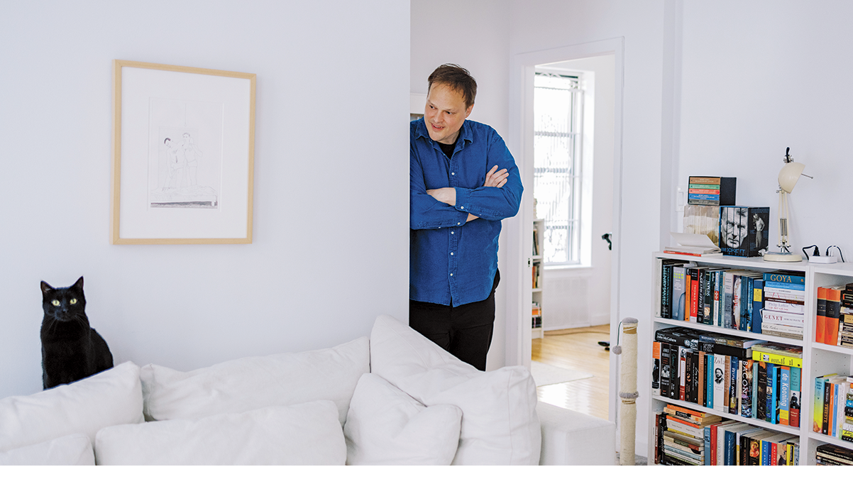 Indoor photograph of a man in a bright blue looking into a room at a black cat on a white sofa