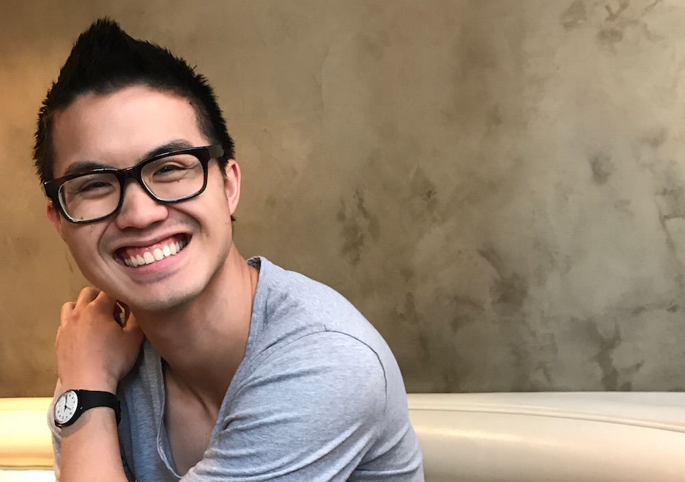 Joshua Nguyen smiles at the camera wearing a gray t-shirt and dark rimmed glasses with short dark hair.