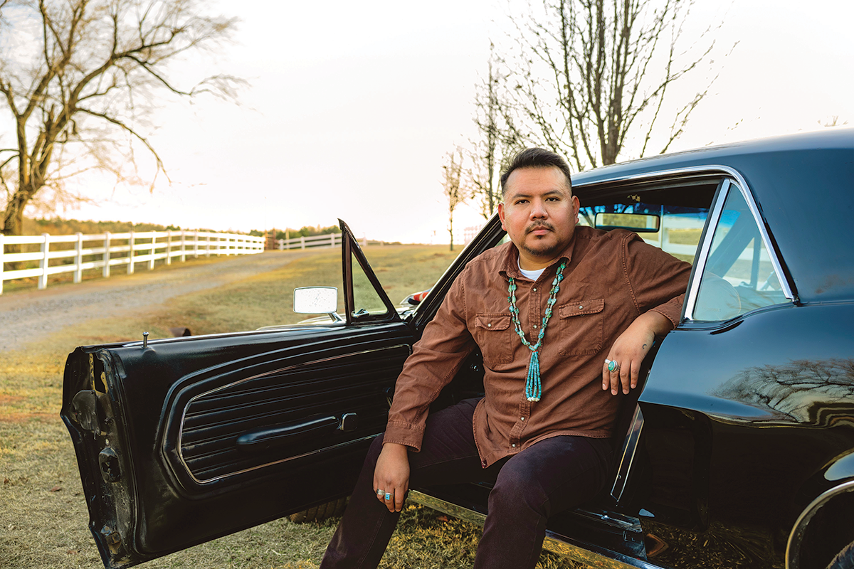 Outdoor photograph of a man with short dark hair and turquoise jewelry sitting on the edge of the front seat of a Mustang with an open door