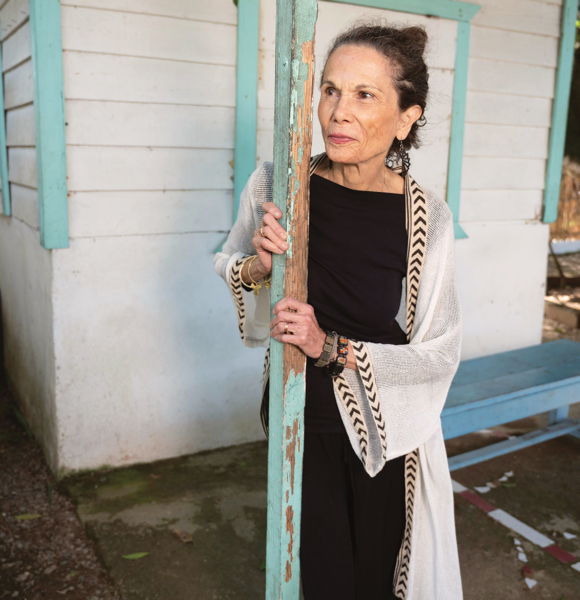 Outdoor photo portrait of a woman with hair pulled back, dark clothes, and a long tan sweater standing behind a weathered wooden beam and in front of a white and teal building