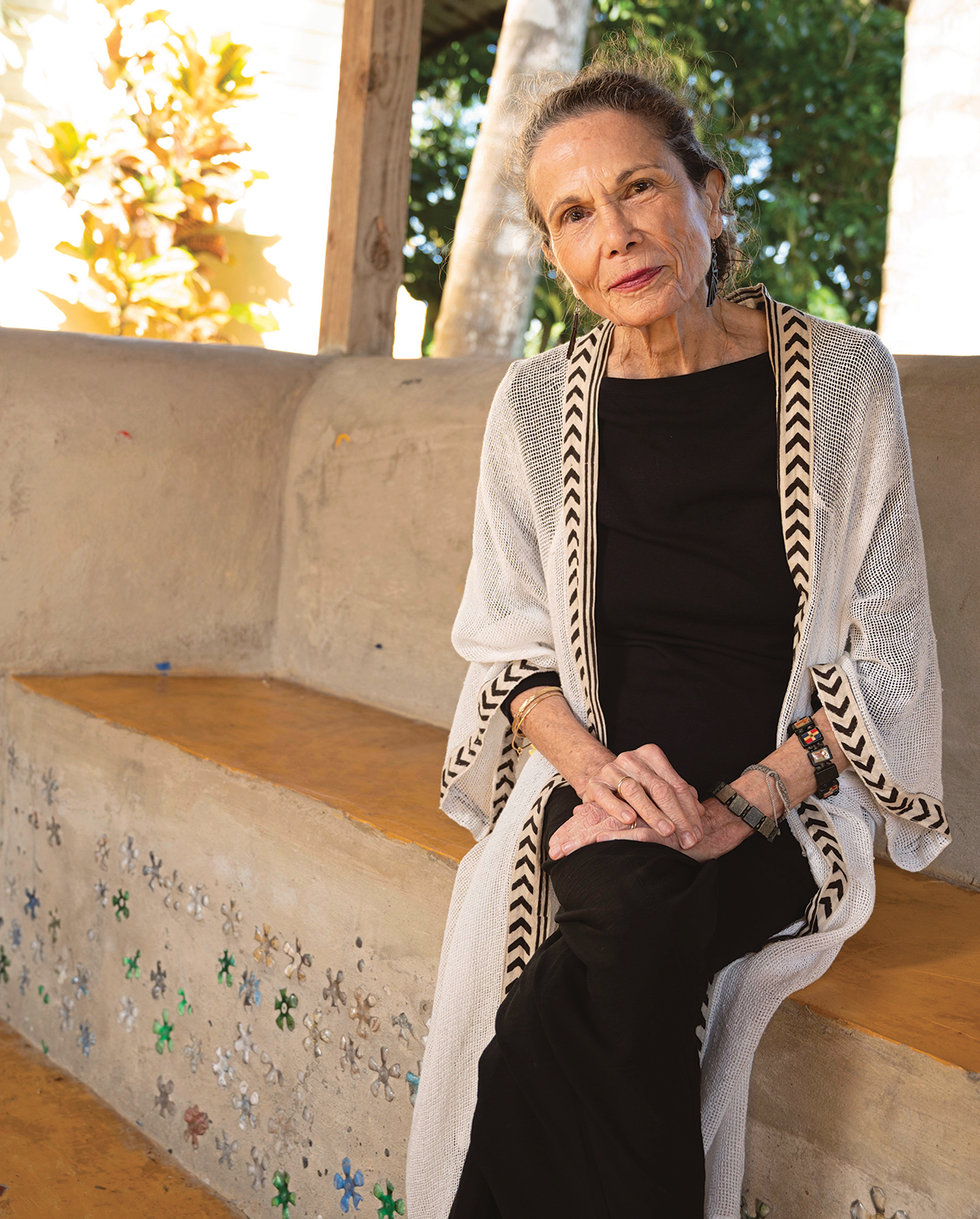 Outdoor photo portrait of a woman in black clothing and a long tan sweater with a chevron design at the edges sitting on a stone bench in a tropical environment 