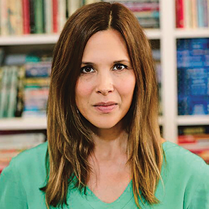 Indoor photo portrait of a woman with shoulder-length brown hair and a green sweater in front of bookshelves