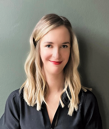 A photo portrait of Katie Greenstreet, a white woman with long slightly wavy blonde hair and blue eyes. She wears a black blouse and red lipstick, and is photographed against a plain dark background.