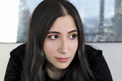 A photo portrait of Mina Hamedi, a Middle Eastern woman with light olive skin and brown hair. She wears a touch of bright green and coral eyeshadow and looks off to the side.