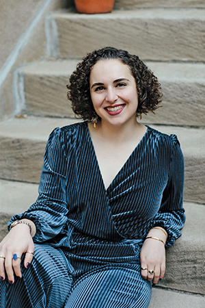 A photo portrait of Natalie Edwards, a woman with light skin and dark brown curly hair. She smiles brightly while leaning one arm against a stair. She wears a dark velvet jumpsuit, a dark lipstick, and assorted jewelry.