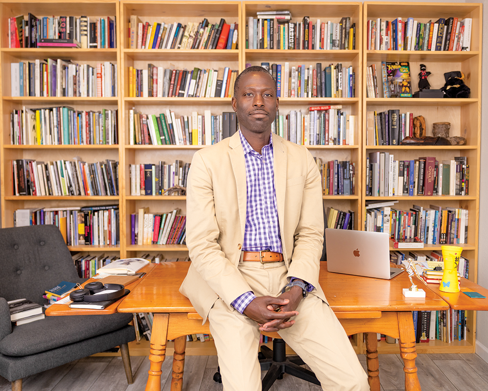 Indoor photo portrait of a Black man in a tan suit leaning back against a table in front of full bookshelves 