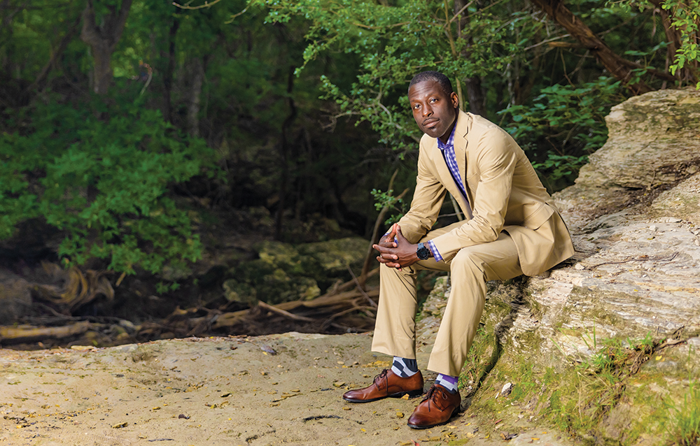 Outdoor photograph of a Black man wearing a tan suit and sitting on an outcrop in a wooded area