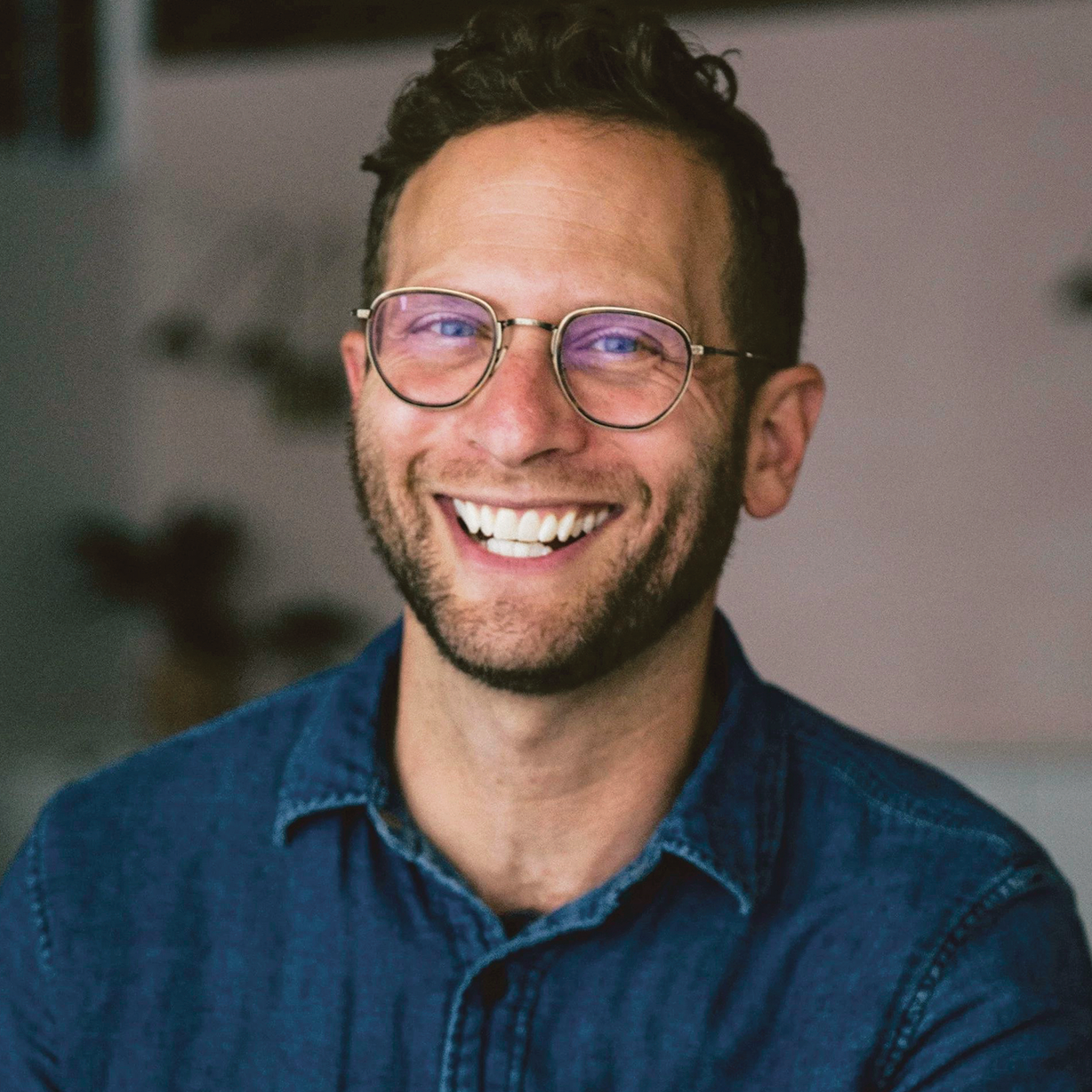 Indoor photo portrait of a man with short wavy hair, wire-rimmed glasses, and a blue shirt