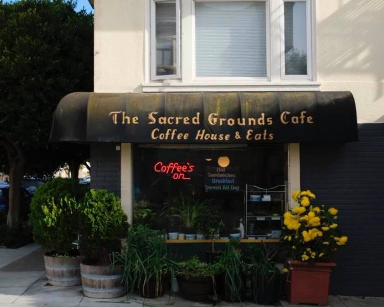 Storefront of Sacred Grounds Cafe with dark awning and plants outdoors.