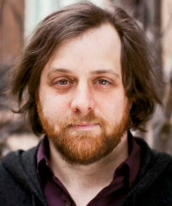 A bust portrait of Craig Morgan Teicher, a white man with medium-brown hair and beard. He wears a dark purple polo and a black zip-up hoodie. The background is softly blurred.