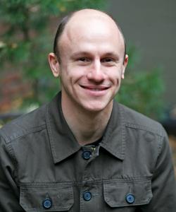 A photo portrait of Aaron Gilbreath, a white man with very short hair. He wears a brown collared button-up shirt and smiles. He sits in an outdoor setting; the background is blurred.