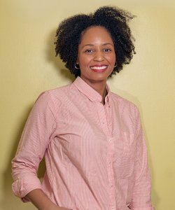 A Black woman with natural hair stands against a chartreuse wall. She wears red lipstick, silver hoop earrings, a collared pink shirt, and a wide smile.