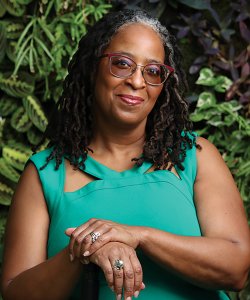 A Black woman with a mobility aid stands against a wall of greenery. She wears tinted glasses and a bright green dress. 