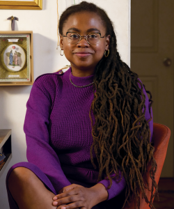 Jamia Wilson, a Black woman with glasses and waist-length locs, sits in a chair. She wears a ribbed purple dress and smiles slightly. Jamia Wilson