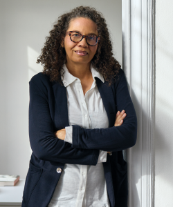 Tanya McKinnon, a Black woman with glasses wearing a navy blue blazer with a white collared shirt, leans against a doorframe.
