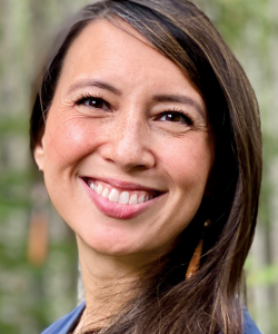A photo portrait of Annie Wenstrup, an Indigenous woman with medium skin, brown hair, and a bright smile. She wears a blue shirt and has her hair parted on the right side.