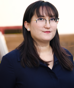Amanda Orozco - credit Kailena Mai A photo portrait of Amanda Orozco, a woman with light skin and long, dark brown hair with bangs. She wears a navy blue button-up blouse and a neat red lip with silver and pearl earrings. The background is blurred.