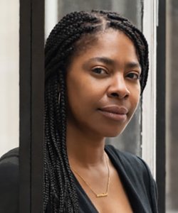 Portrait of a woman with long braided hair wearing a gold necklace and a black sweater over a black shirt. 