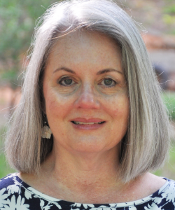 A white woman with gray shoulder-length hair gazes forward with a slight smile. She wears a blue and white floral top against a natural background.