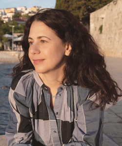 Outdoor photo portrait of a woman with dark curly hair and blue patchwork blouse leaning on a short wall and and gazing off to the side