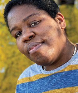 A Black person tilts their head with a neutral expression. They wear a silver chain and blue-and-yellow-striped shirt and stand in a background of greenery.