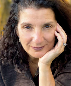 A photo portrait of Carla Panciera, a middle-aged white woman with brown curly hair. She wears a brown jacket and brown lipstick and leans on one hand.