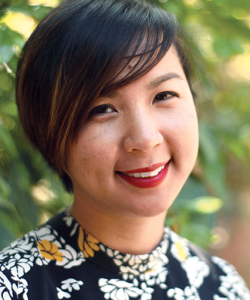 Cathy Linh Che, an Asian woman with short black hair parted to the left side of her head, smiles. She wears a black patterned blouse and red lipstick.