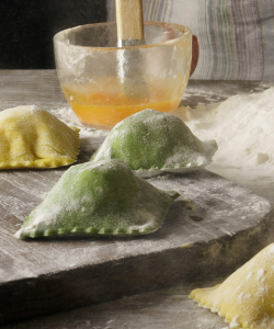 Flour plump homemade ravioli sit on the kitchen table. They are lightly dusted with flour, waiting to be cooked. Toward the back is a small glass bowl with an egg mixture and a mound of white flour.