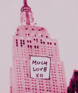 Edited photo of the upper half of the Empire State Building. Everything is in pink tones. A small white square with the handwritten words “MUCH LOVE XO” is pasted on top of the photo, on the face of the building.
