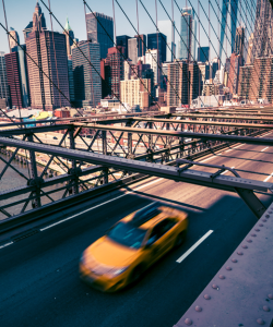 A solitary yellow taxicab crosses over the Brooklyn Bridge on a clear, bright day. Skyscrapers in downtown Manhattan are visible in the background. 