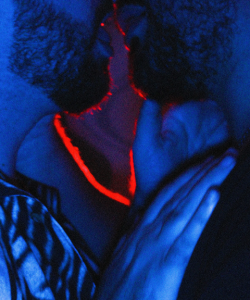 Close-up of two men with scruffy facial hair embracing in a dark room. A red light behind them illuminates the edges of their skin.