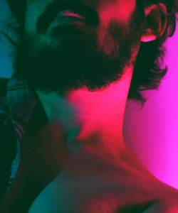 Close-up of a man with a dark, full beard and mustache. The photo is cropped just above his mustache and just below his chest. He is lit by purple lighting on one side and blue on the other. 