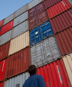 A person with their back to the camera gazes up at a stack of large shipping containers 