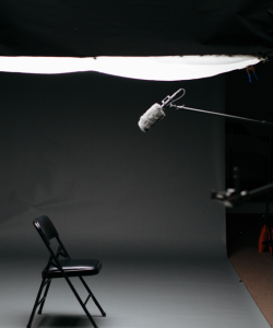  View of a set for recording an interview. A single black fold-out chair sits on a slate gray paper floor and backdrop. A microphone at the end of a boom floats above the chair, and a bright overhead light illuminates the scene.