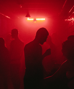 Various small groups of people mill around in a dark club, silhouetted by red lighting, which also causes the tile wall on the right-hand side to shimmer.