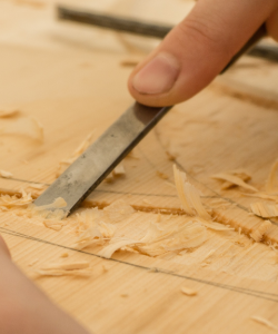 Close-up of a person's hand using a wood chisel to cut a pattern into a light blond wood. Small, thin shavings of wood are scattered around the area being cut.