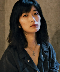 A photo portrait of Cleo Qian, a young East Asian woman. She has shoulder-length black hair and bangs, and wears a button-up corduroy shirt against a neutral background.
