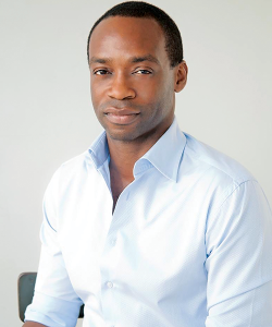 A bust portrait of Rowan Ricardo Phillips, a Black man with very short hair. He wears a light blue collared shirt and a stoic expression.