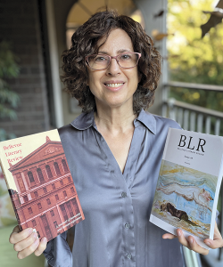 Photo portrait of white woman with blue blouse and curly brown hair standing on outdoor porch holding up two magazines 