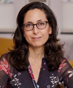 A photo portrait of a white woman with wavy brown hair and glasses smiling slightly. She wears a dark blouse with red florals.